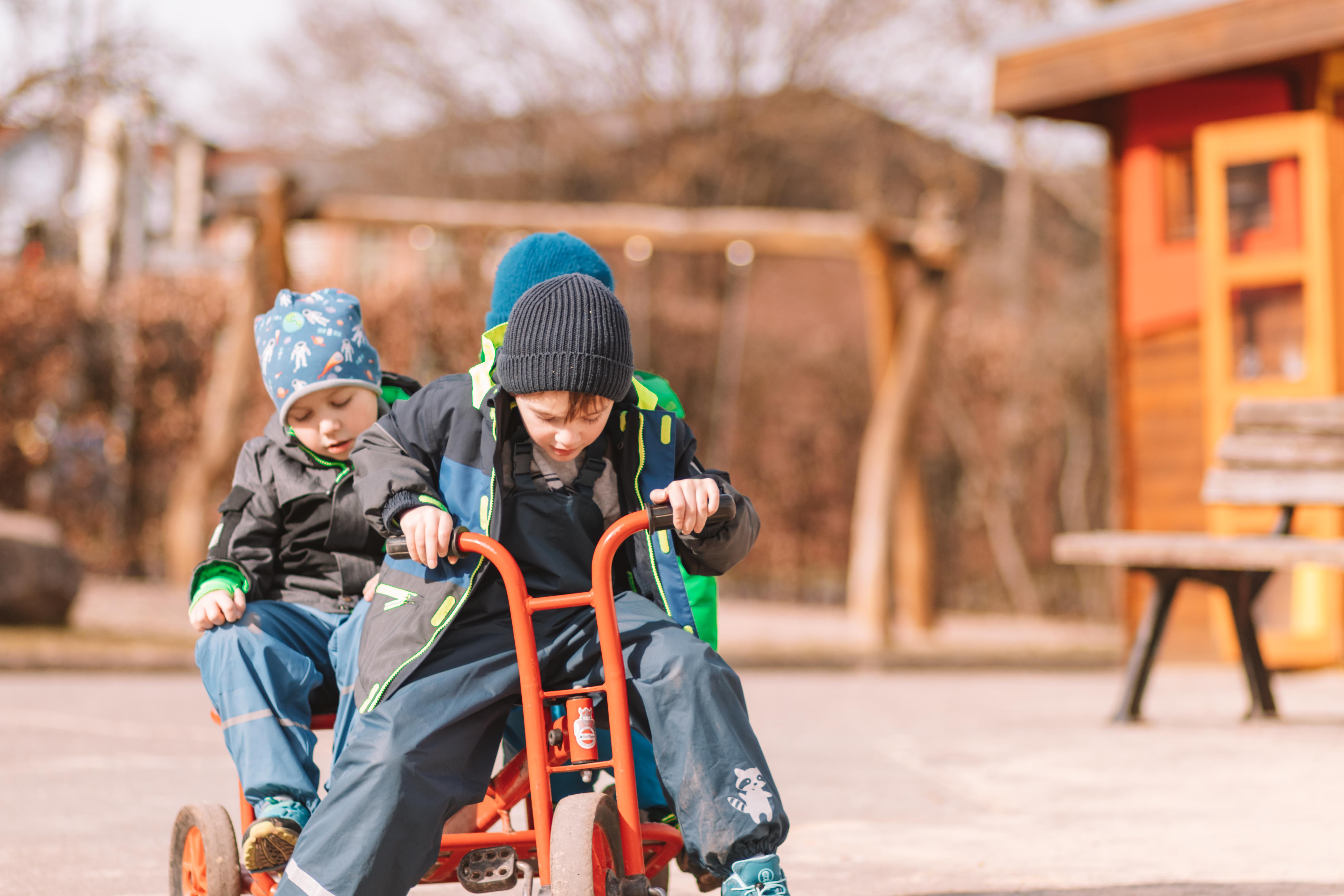 Zwei Kinder spielen fröhlich auf einem roten Tretauto auf einem Spielplatz im Frühling.