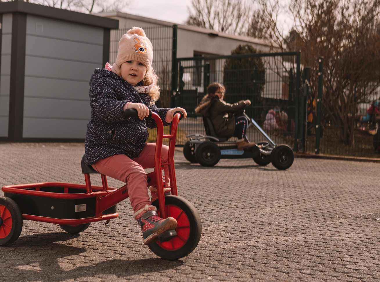 Kleines Mädchen spielt mit einem roten Dreiradwagen im Freien.